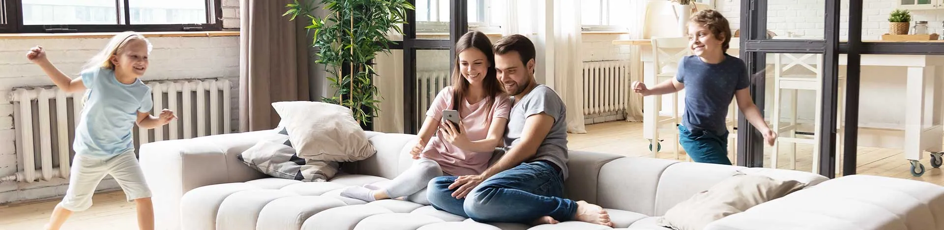 Wohnmanufaktur - Zwei Erwachsene sitzen gluecklich auf dem Sofa und zwei Kinder toben in der Naehe Zwei Erwachsene sitzen glücklich auf dem Sofa und zwei Kinder toben in der Nähe